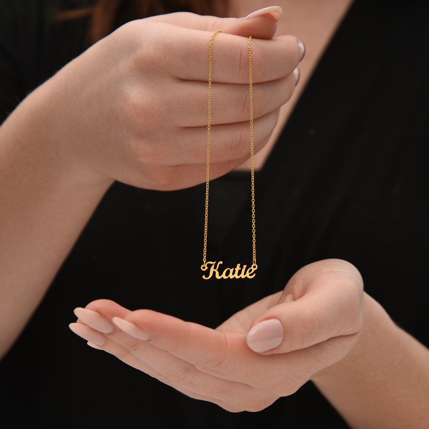 A person's hand holding a gold necklace with the name "Katie" engraved on it.