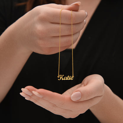 A person's hand holding a gold necklace with the name "Katie" engraved on it.