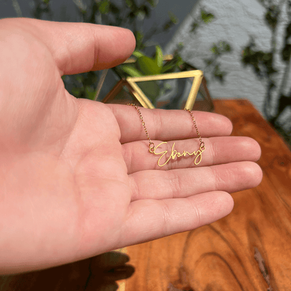 A hand holding a gold necklace with the name "Ebony" engraved on it.